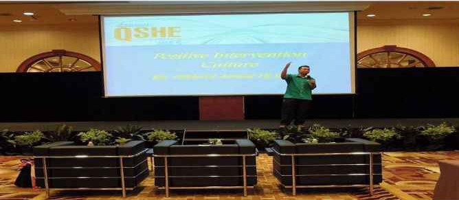 Speaker on a hotel ballroom stage in front of a QSHE conference screen